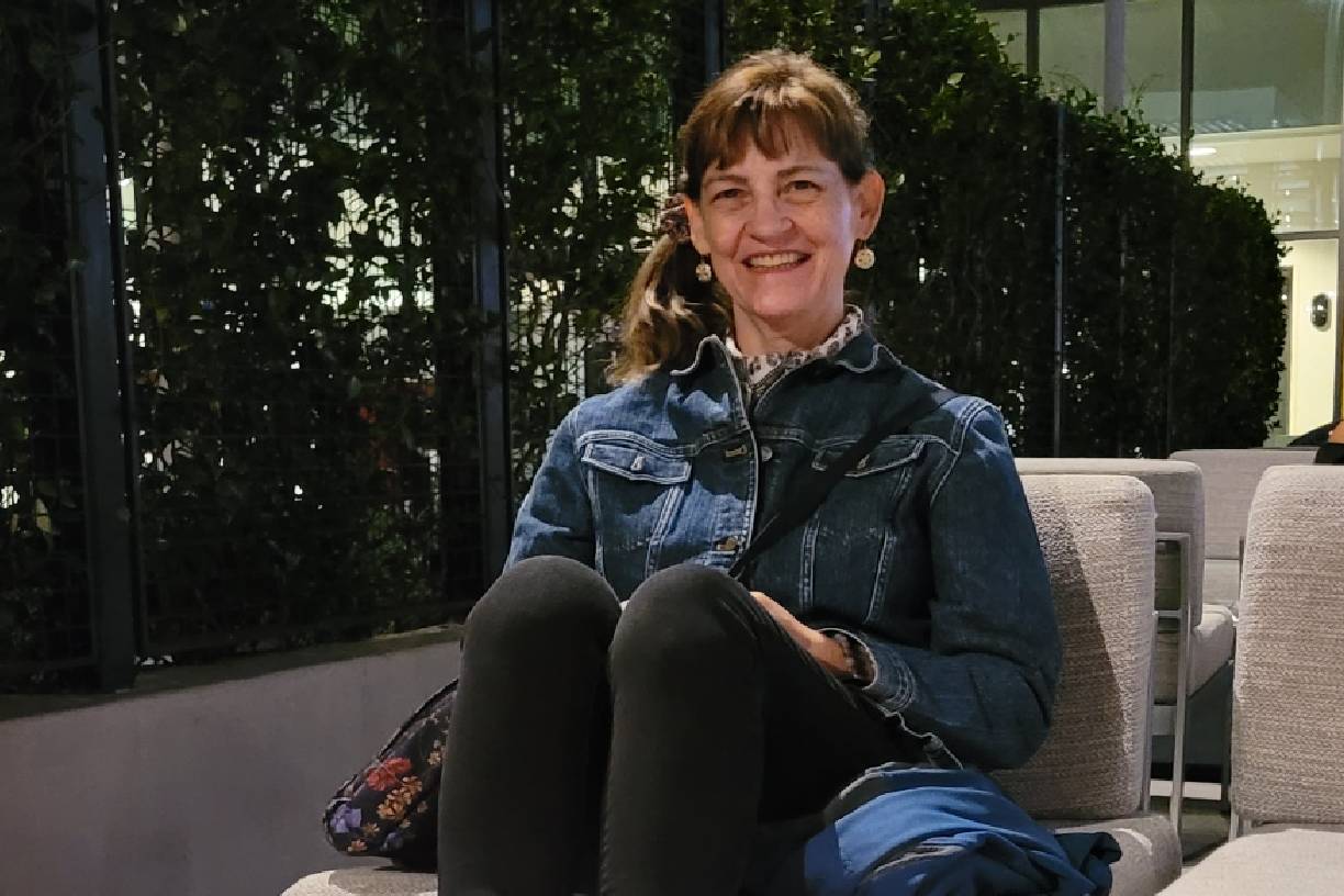 Smiling woman sitting outdoors in casual denim jacket and black pants.